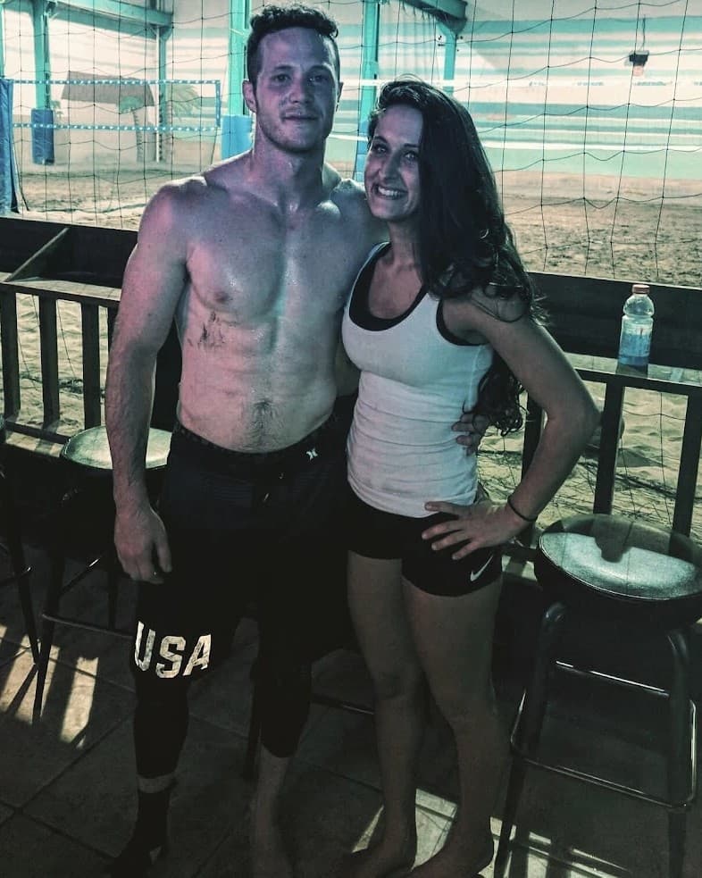 Sheldon and Danielle standing together beside an indoor sand volleyball court at Hot Shots, a net and sand behind them