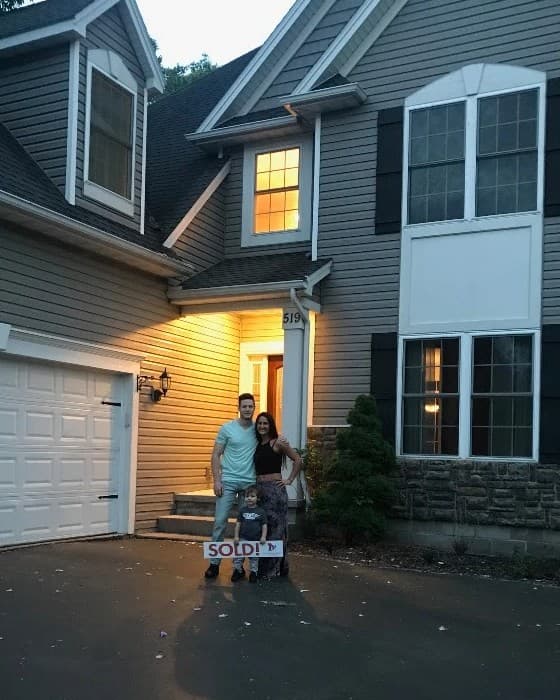 Sheldon and Danielle standing at the front door of their new house at dusk with a small boy, holding a sold sign